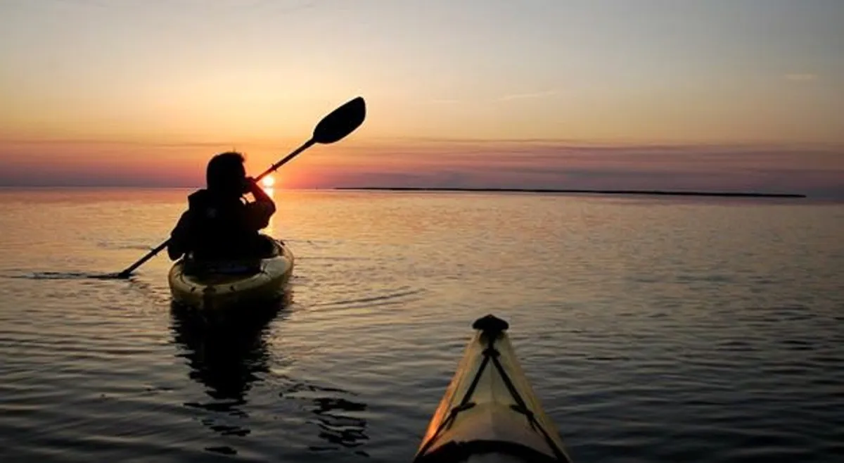 A serene view scene capturing a kayaker gliding across calm water at sunset, with glowing skies reflecting in the Currituck Sound. The atmosphere is tranquil and awe-inspiring, speaking to the natural beauty guests will witness from their balconies.