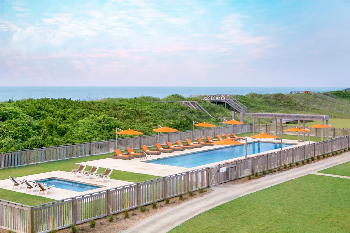An inviting resort pool area with comfortable lounge chairs, orange umbrellas, lush greenery, and an expansive view of the ocean beyond. The setting suggests relaxation, socializing, and easy access between pool and beach environments.