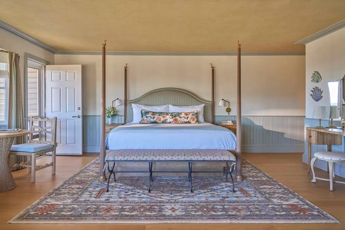 A spacious bedroom featuring a four-poster king-size bed with luxurious linens, a decorative bench at the foot of the bed, soft area rug, and large windows that flood the room with natural light. The mood is cozy and serene, inviting restful sleep or a relaxing morning routine.