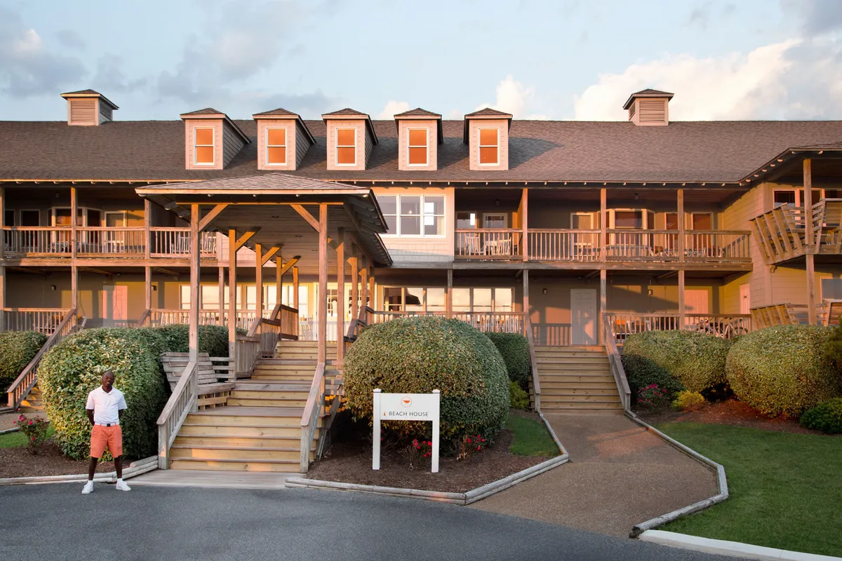 The exterior of the property, featuring a welcoming entrance with stairs, a beach house sign, round bushes, and a figure standing at the doorway. The stairs are clearly visible, which relates to the accessibility note, while the overall mood is inviting and relaxed.