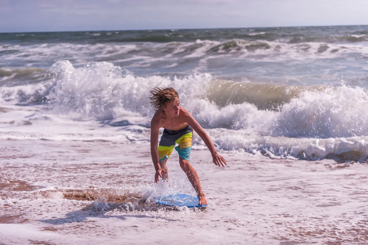 A sunny beach scene featuring a boy surfing in the waves, with the expansive shoreline in the background. The image radiates joy and highlights active, playful moments by the ocean.