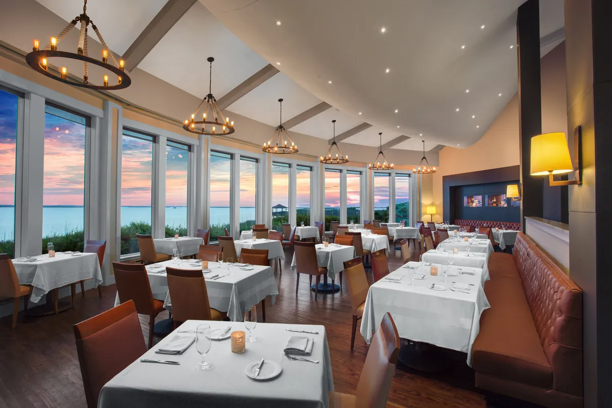A sophisticated interior dining space with large windows framing a breathtaking sunset, elegantly set tables, and sparkling chandeliers. The refined ambiance suggests special dining experiences with a view of the water.