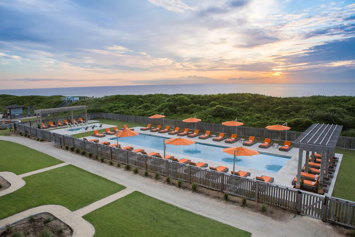 This exterior image captures a serene swimming pool area lined with vibrant orange lounge chairs, set against the dramatic backdrop of a sunset over the Atlantic Ocean. The tranquil water, inviting seating, and the expansive ocean view create an atmosphere of relaxation and luxury.