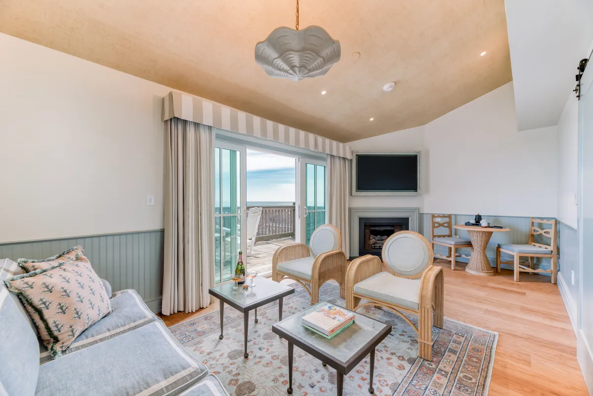 The image showcases a serene, sunlit living area with expansive windows that offer sweeping ocean views. The room features a stylish light fixture, plush and inviting seating, and elegant coffee tables, all arranged to encourage relaxation and conversation. The natural light floods the space, and the tranquil scene hints at an inviting balcony just beyond the glass, perfect for enjoying sunrises or sunsets over the water.
