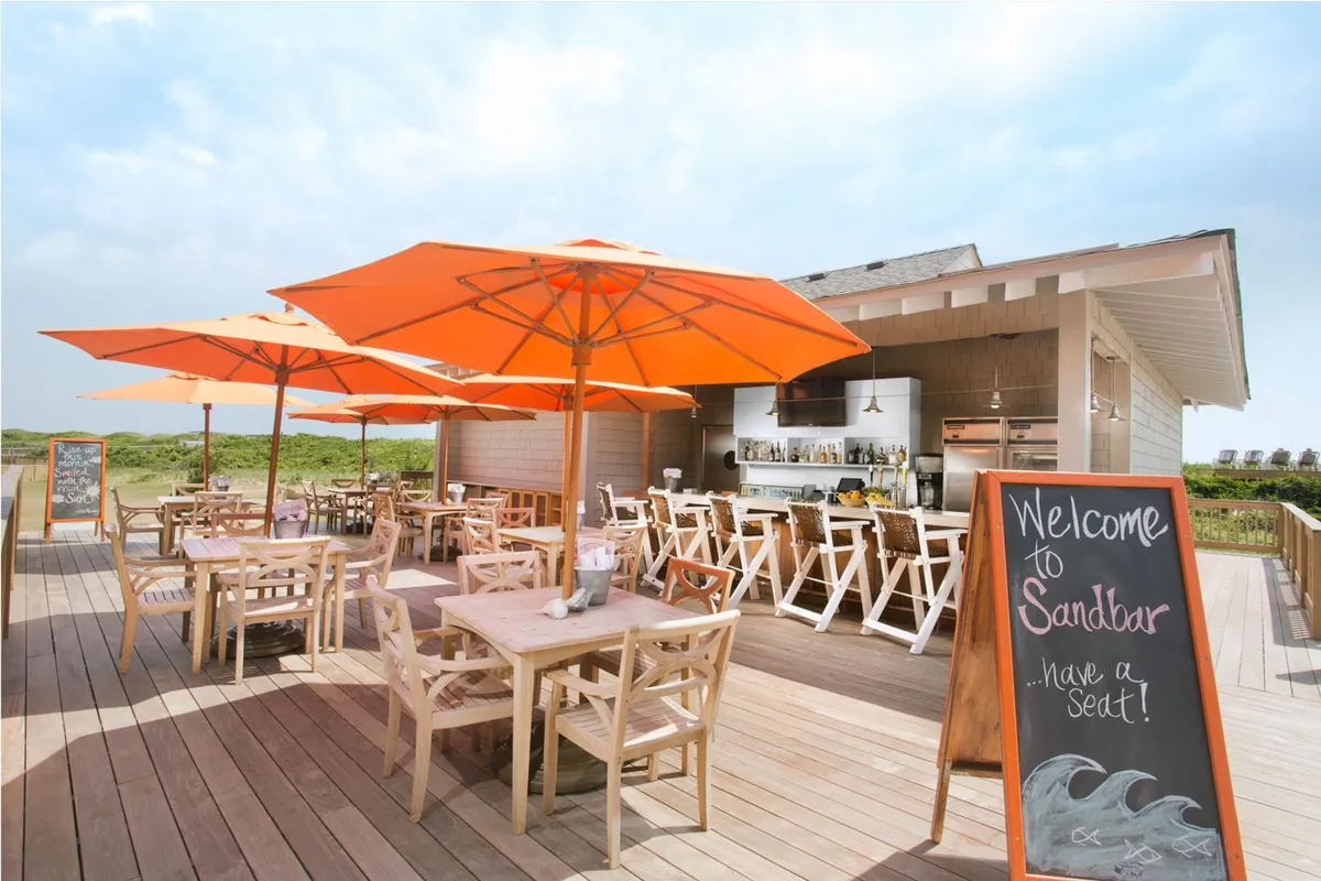 An exterior dining area shaded by bright orange umbrellas, with wooden tables and a visible bar area. The relaxed setting invites guests to enjoy alfresco dining and social gatherings in the fresh coastal air.