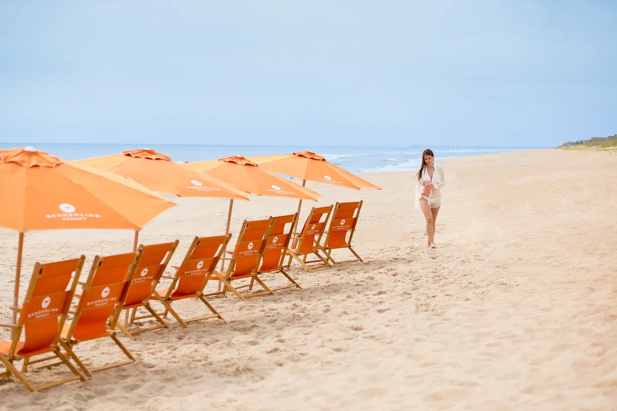 A coastal scene featuring beach chairs and umbrellas set along the sandy shoreline, with the ocean stretching into the horizon and a woman strolling by the water. The tranquil setting is tailored for effortless beach days.