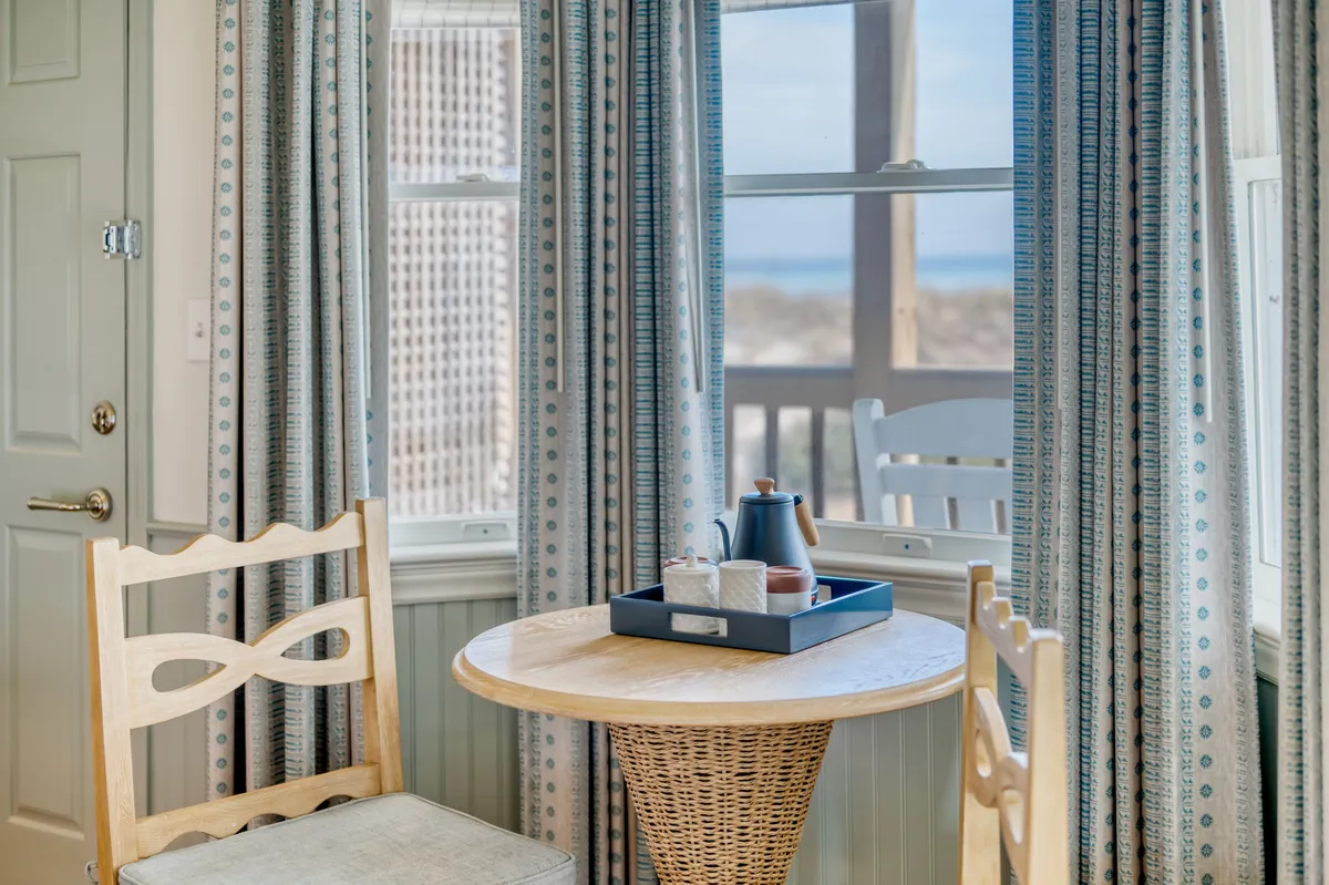 A stylish living and dining area with a round wooden table, woven chair, a tray with teapot and cups, and blue-patterned curtains. The atmosphere is relaxed and comfortable, ideal for intimate mornings or restful evenings.