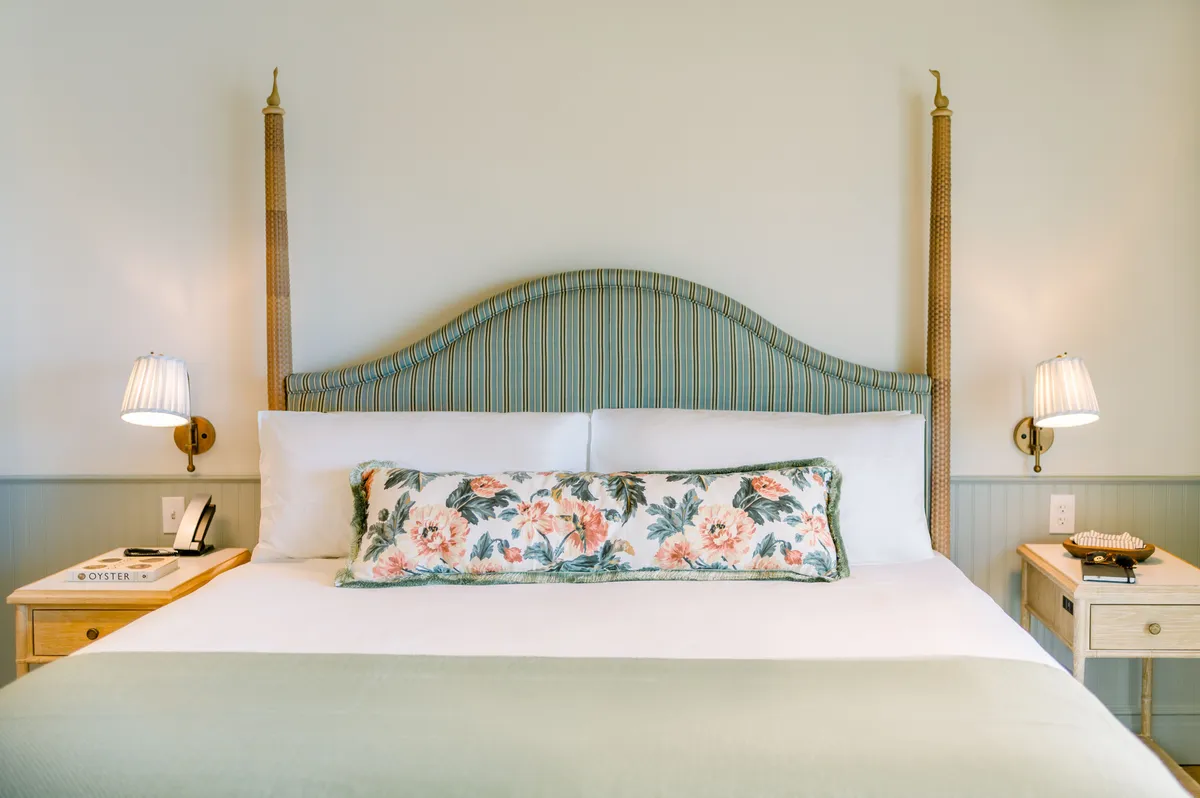 This image features a bed with a patterned headboard and a floral accent pillow, accompanied by a nightstand and lamp. Natural light filters into the room, creating a serene and comfortable atmosphere, suggestive of a restful retreat perfect for all seasons. The setting evokes relaxation, warmth, and the comfort of snuggling under cozy blankets.