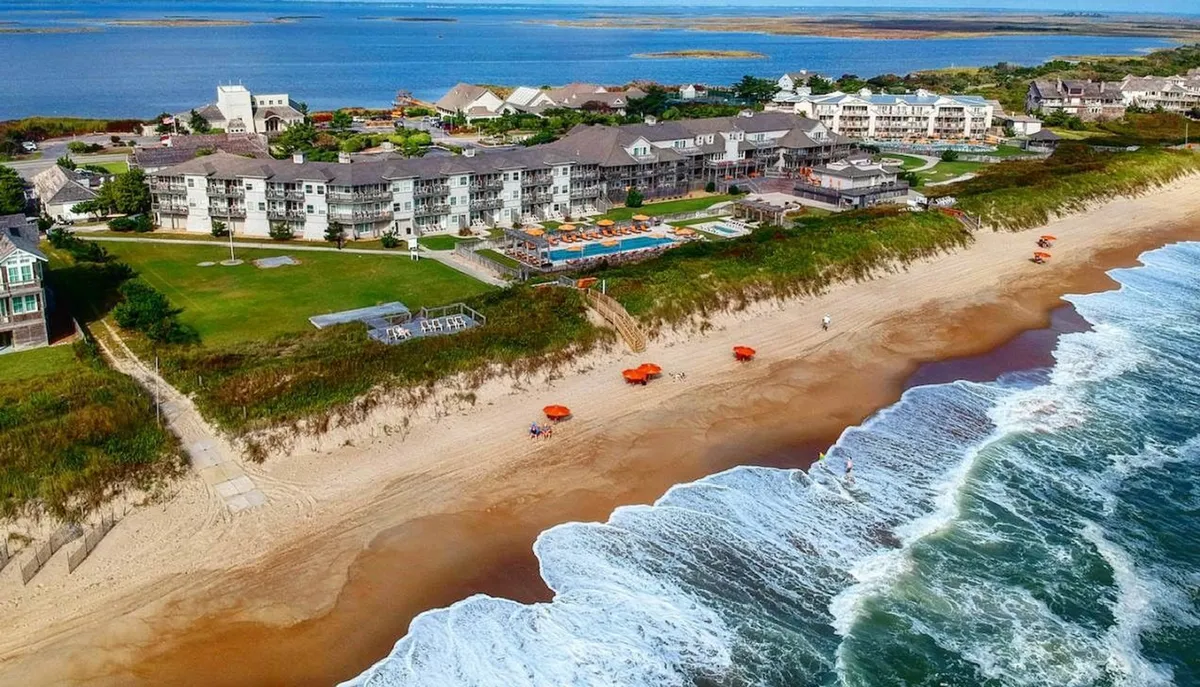 A wide exterior view capturing both the sandy beach and ocean in the background, the hotel pool lined with umbrellas, green landscaping, and spaces where families can gather. There is vibrant beach activity and a sense of access to both the water and recreational amenities.